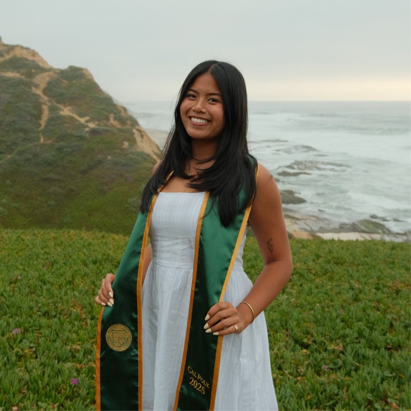 A picture of Jasmine standing in a graduation outfit wearing a Cal Poly green stahl. The picture has the ocean in the background and some rolling green hills in the forground.