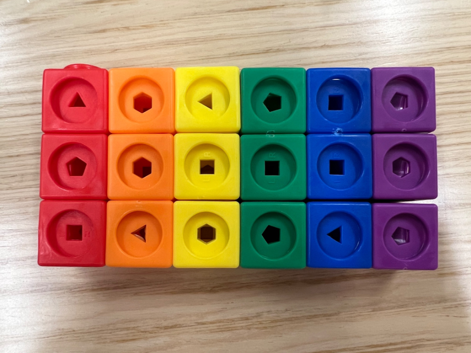 A picture of a set of Mathlink blocks on a desk. The blocks are connected together forming three rows and six columns. The columns are red, orange, yellow, green, blue, and purple. Each block within a row and colum has a shape in the center (e.g., triangle, square hexagon).