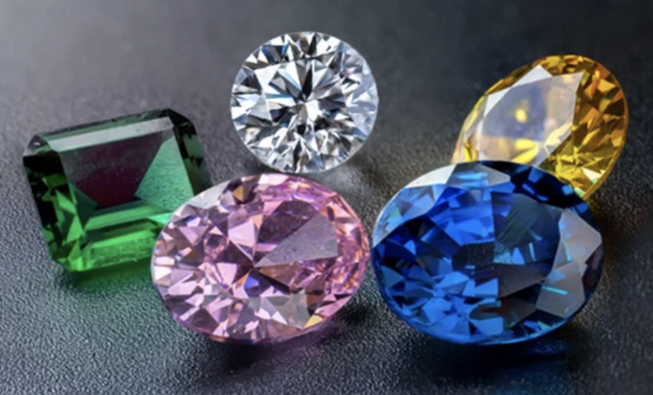 The image displays five colorful gemstones placed on a dark surface. Each gem has a distinct color and shape. A rectangular green gem (emerald-like) on the left. A round white gem (resembling a diamond) at the top. An oval pink gem in the center. An oval blue gem in the foreground. An oval yellow gem in the background on the right. The gems are cut and polished, reflecting light and showing their intricate facets. They appear to represent a variety of precious stones with vibrant colors.