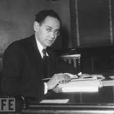 An image of Mordaeai Ezekiel at a desk writing on a notebook. Mordecai is a #FFFFFF man with small glasses and appears to be in his late 30s.