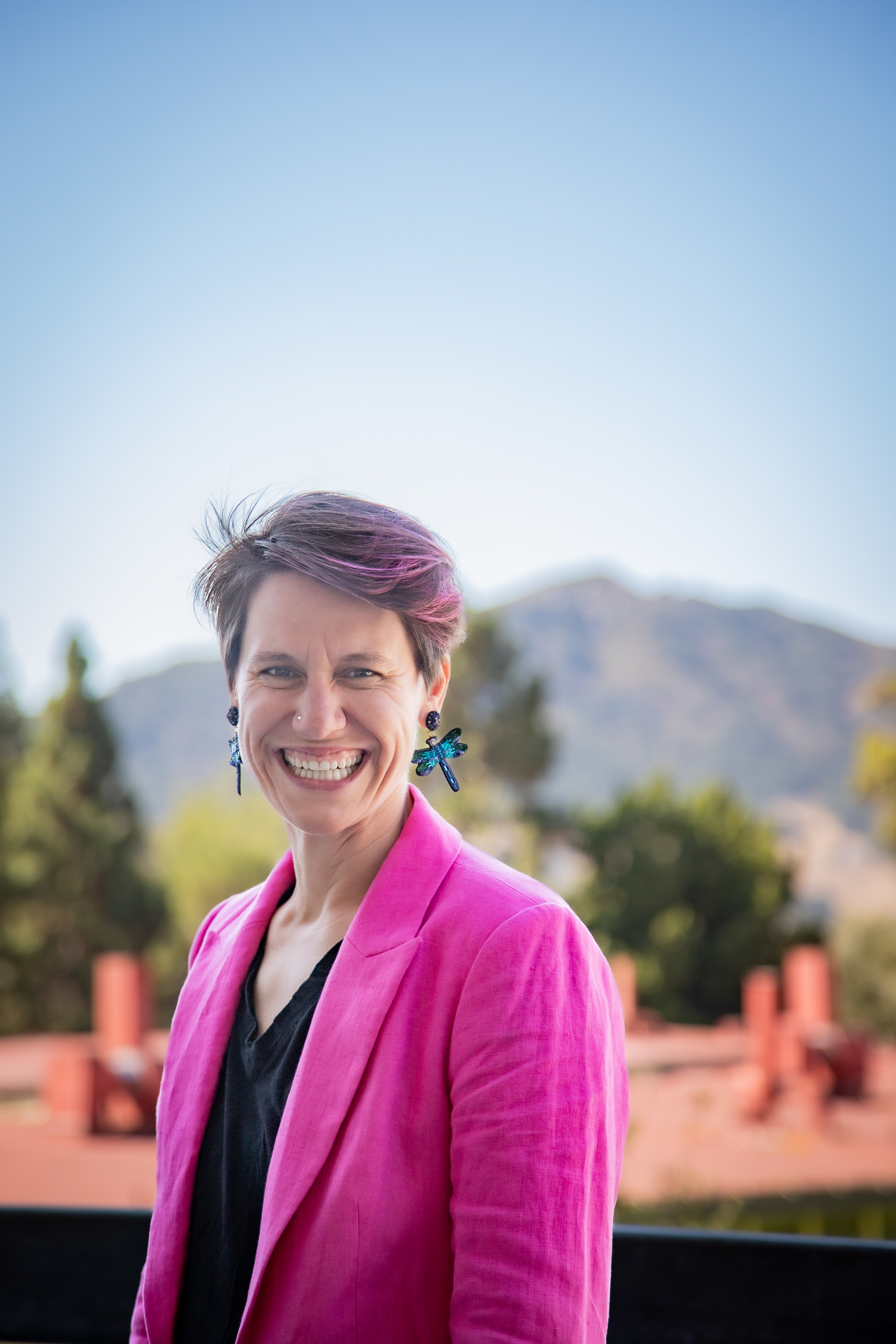 A headshot of Dr. Theobold, in a pink blazer at the top floor of Building 25, overlooking Bishop Peak