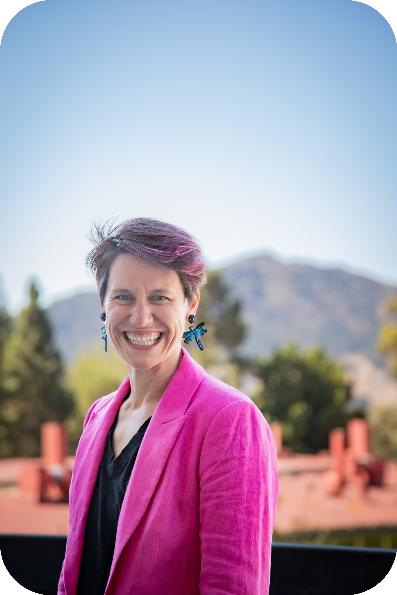 A headshot of Allison, a non-binary identifying person with short brown hair and fun patterned glasses. They are wearing a pink blazer, with the green mountains of San Luis Obispo in the background.