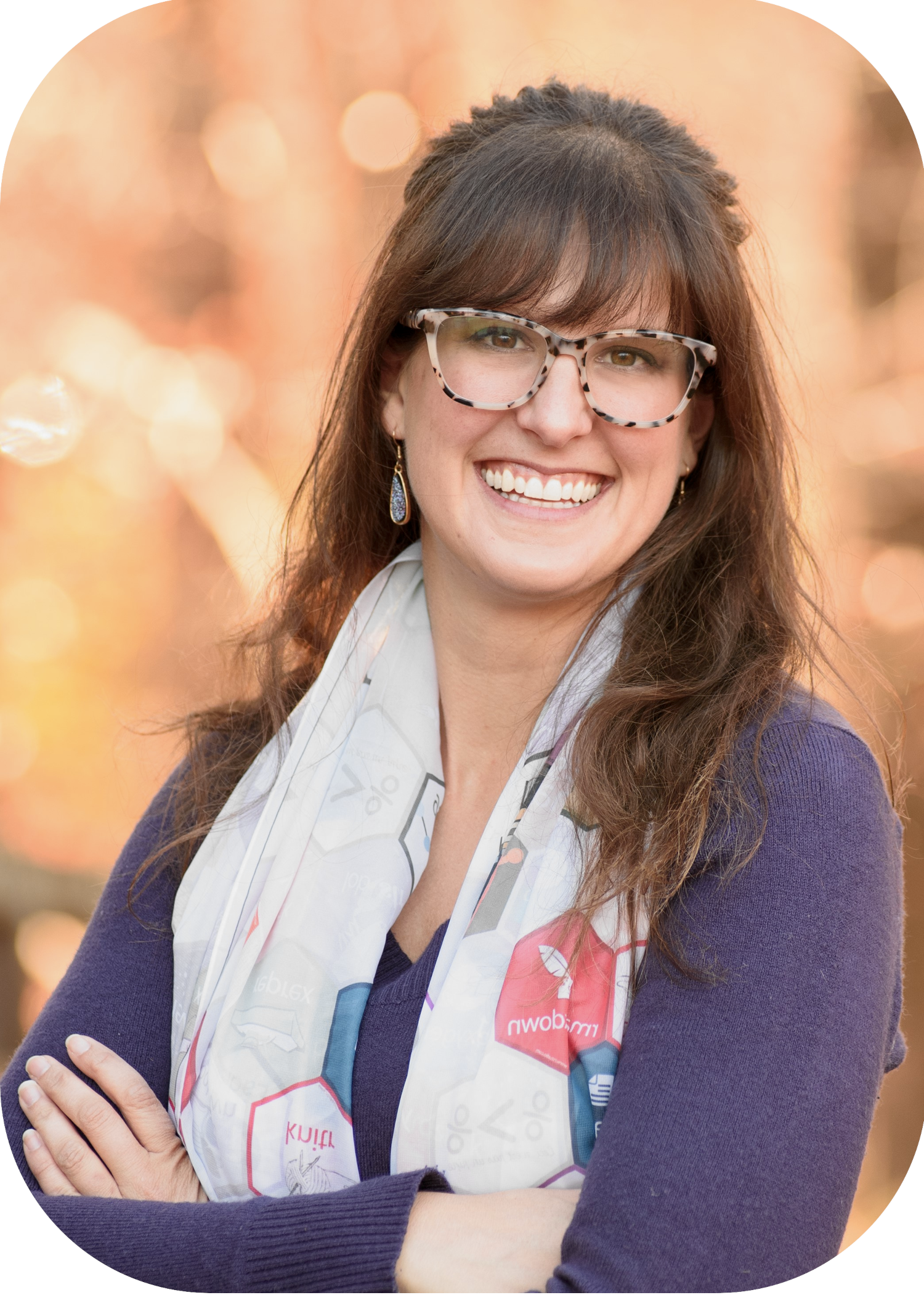 A headshot of Kelly, a woman with with long brown hair and bangs. Kelly smiles warmly while standing outdoors against a soft background of autumn tones. She wears tortoiseshell glasses, a purple sweater, a tidyverse-themed scarf, and dangling blue earrings, with arms confidently crossed.