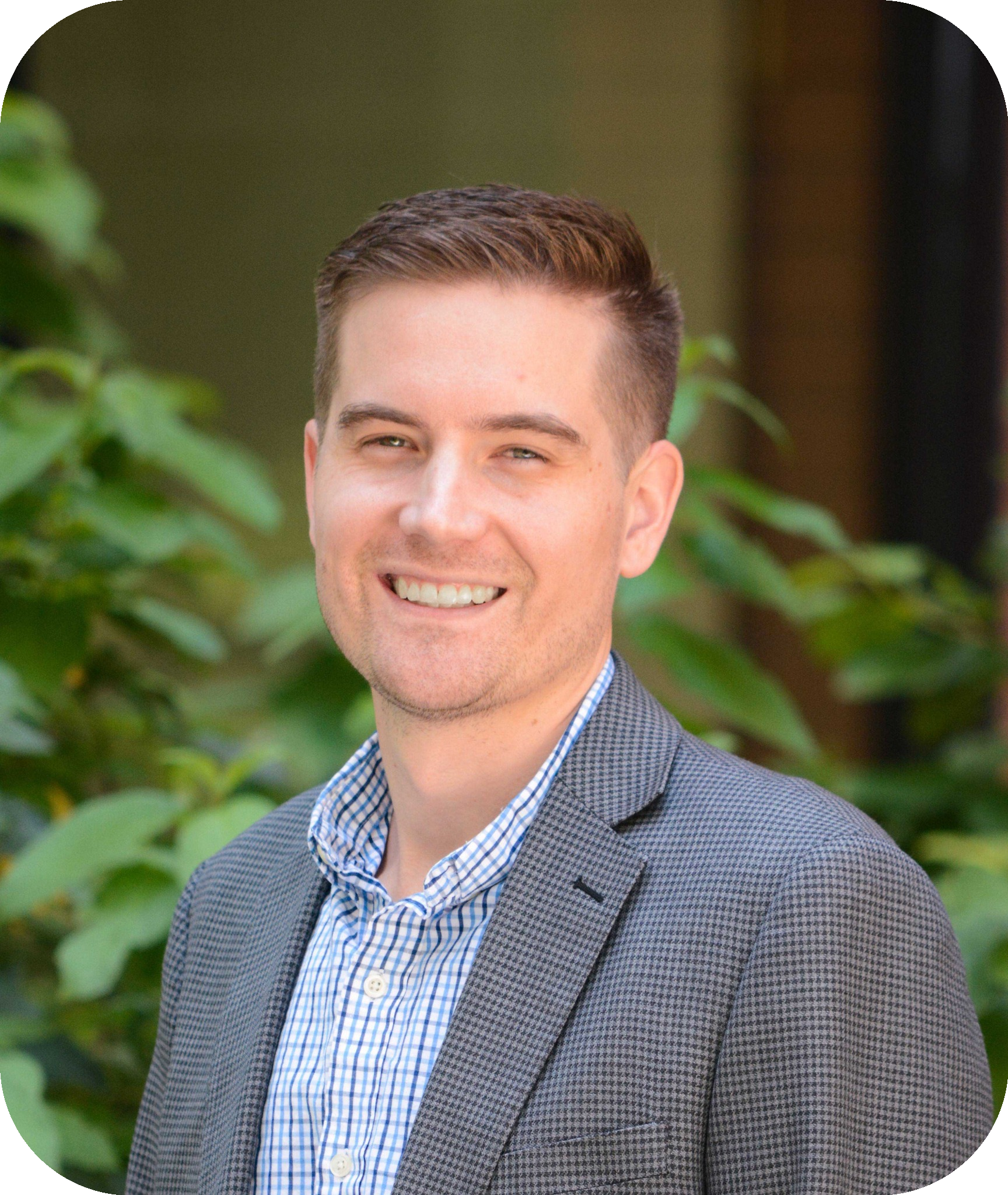 A headshot of Tyson, a man with short light brown hair. Tyson smiles confidently while standing in front of greenery and a blurred building background. He is wearing a grey patterned blazer over a blue and white checkered shirt, giving a professional and approachable appearance.